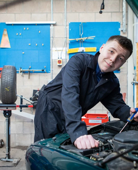 Student in a black overall smiling while working on a car engine, with a workshop setup in the background. Student in a black overall smiling while working on a car engine, with a workshop setup in the background.